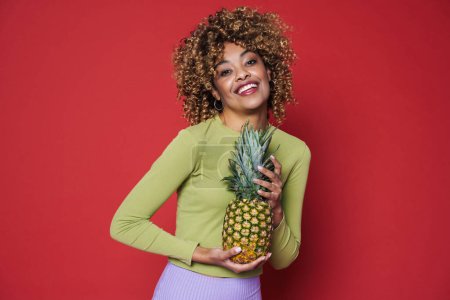 Foto de Young black woman laughing while posing with pineapple isolated over red background - Imagen libre de derechos
