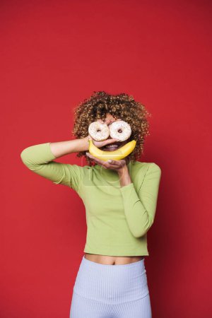 Foto de Young black woman with afro curls having fun holding donuts and banana isolated over red background - Imagen libre de derechos
