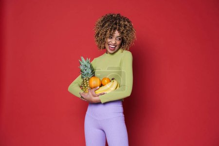 Photo for Young black woman laughing while posing with fruits isolated over red background - Royalty Free Image