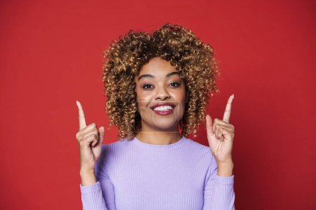 Foto de Young black woman with afro curls smiling and pointing at copyspace isolated over red background - Imagen libre de derechos
