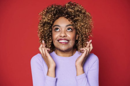 Téléchargez les photos : Young black woman with afro curls smiling and holding fingers crossed isolated over red background - en image libre de droit
