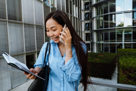 Téléchargez les photos : Asian brunette woman talking on mobile phone while standing with planner outdoors - en image libre de droit