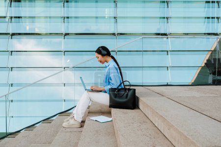 Téléchargez les photos : Asian brunette woman in headphones working with laptop while sitting on stairs outdoors - en image libre de droit