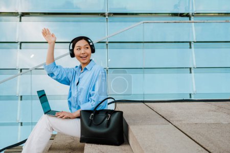 Téléchargez les photos : Asian brunette woman in headphones working with laptop while sitting on stairs outdoors - en image libre de droit