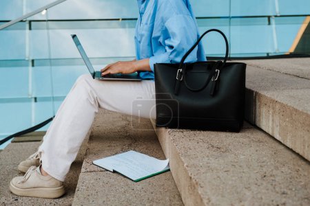 Photo for Young woman working with laptop while sitting on stairs outdoors - Royalty Free Image