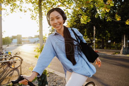 Photo for Asian woman in headphones smiling while riding bicycle on city street - Royalty Free Image