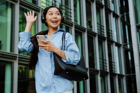 Téléchargez les photos : Asian brunette woman in headphones using cellphone while walking on city street - en image libre de droit