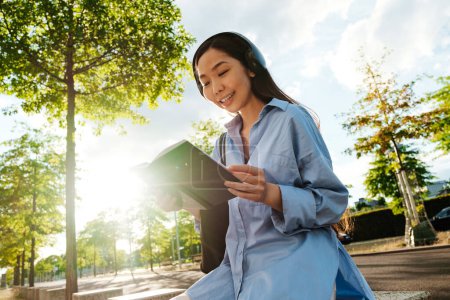 Téléchargez les photos : Asian brunette woman in headphones reading book while sitting on bench outdoors - en image libre de droit