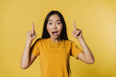 Photo for Young asian woman expressing surprise while pointing fingers upward isolated over yellow background - Royalty Free Image
