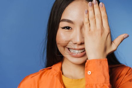 Téléchargez les photos : Young asian woman wearing t-shirt smiling while covering her eye isolated over blue background - en image libre de droit