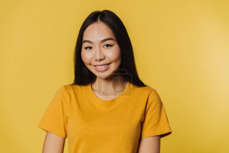 Téléchargez les photos : Young asian woman wearing t-shirt smiling while looking at camera isolated over yellow background - en image libre de droit