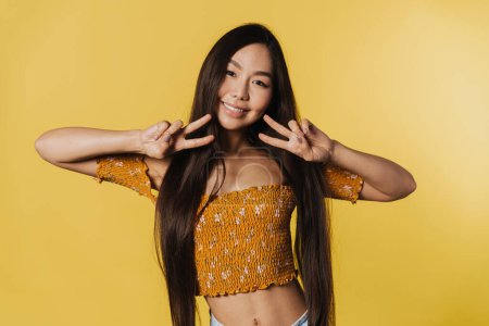 Foto de Young asian woman wearing top smiling and gesturing at camera isolated over yellow background - Imagen libre de derechos