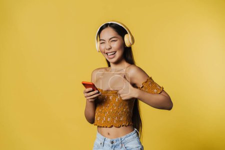 Foto de Young asian woman pointing finger at her cellphone while listening music isolated over yellow background - Imagen libre de derechos