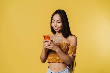 Téléchargez les photos : Young asian woman wearing top smiling and using cellphone isolated over yellow background - en image libre de droit