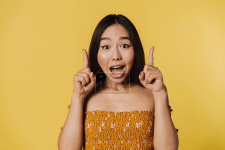 Photo for Asian woman expressing surprise while pointing fingers upward isolated over yellow background - Royalty Free Image