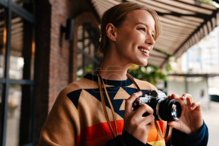 Téléchargez les photos : Joyeux photographe femme blonde portant un pull prenant des photos sur un appareil photo vintage tout en marchant à l'extérieur - en image libre de droit
