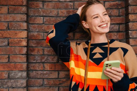 Foto de Mujer joven sonriente en suéter colorido usando teléfono inteligente mientras está de pie sobre la pared de ladrillo en la calle de la ciudad - Imagen libre de derechos
