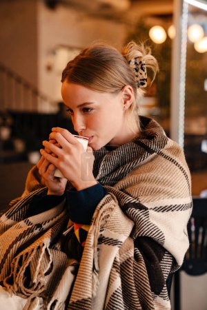 Photo for Young white woman wrapped in blanket drinking coffee at cafe indoors - Royalty Free Image