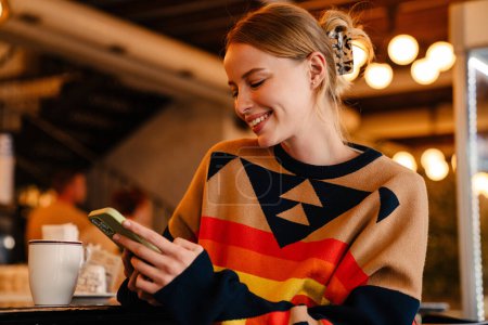 Photo for Young white woman in sweater smiling and using cellphone at cafe indoors - Royalty Free Image