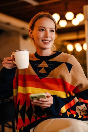Photo for Young white woman in sweater drinking coffee and using cellphone at cafe indoors - Royalty Free Image