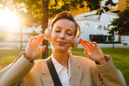 Foto de Retrato de una joven hermosa mujer en auriculares con los ojos cerrados disfrutando de la música, mientras que de pie en el césped soleado en la ciudad - Imagen libre de derechos