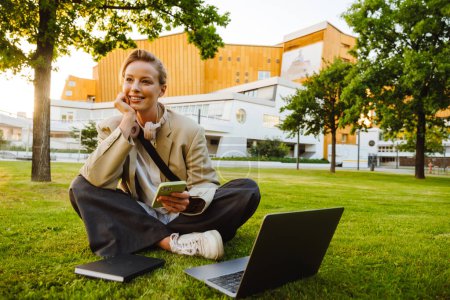 Photo for Young beautiful smiling happy business lady holding phone, propping her head and looking aside, while sitting in lotus pose with laptop and notebook on the lawn in the city - Royalty Free Image