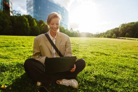 Foto de Young beautiful smiling happy business woman working with laptop, while sitting in lotus pose on sunny lawn in the city - Imagen libre de derechos