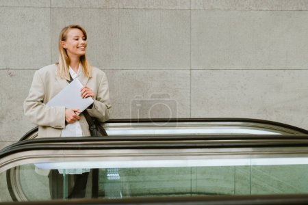 Téléchargez les photos : Jeune belle dame d'affaires souriante en blazer avec ordinateur portable regardant de côté, tout en se tenant debout sur l'escalator - en image libre de droit