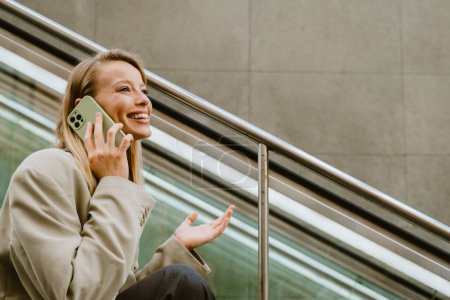 Téléchargez les photos : Jeune belle dame d'affaires souriante heureuse parlant son téléphone gesticulant et regardant de côté, tout en étant assis dans les escaliers à l'extérieur - en image libre de droit