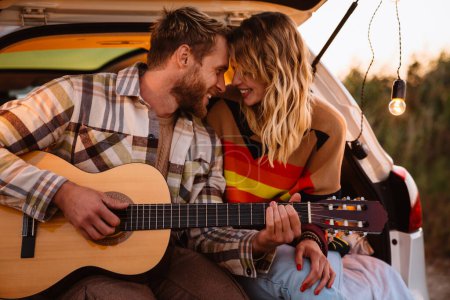 Téléchargez les photos : Joyeux jeune couple blanc souriant et jouant de la guitare assis dans le coffre au bord de la mer au coucher du soleil - en image libre de droit