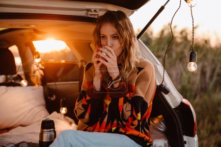 Photo for Happy young white woman wearing sweater drinking tea while sitting in trunk in evening - Royalty Free Image