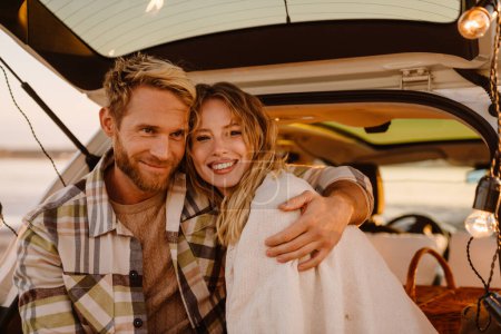 Foto de Happy young white couple smiling and sitting in car trunk together outdoors - Imagen libre de derechos
