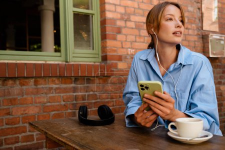 Photo for Young beautiful thoughtful woman with opened mouth in headphones holding phone and looking aside, while sitting with cup of coffee by table near brick wall outdoors - Royalty Free Image