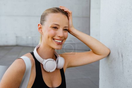 Photo for Young beautiful smiling sporty woman with headphones on her neck propping her head and looking aside, while standing outdoors - Royalty Free Image