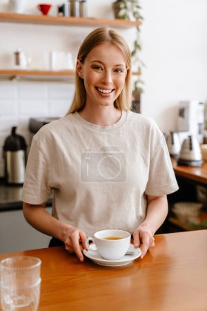 Téléchargez les photos : Young beautiful attractive long-haired smiling woman in t-shirt with cup of tea looking at camera, while standing behind the bar in coffee shop - en image libre de droit