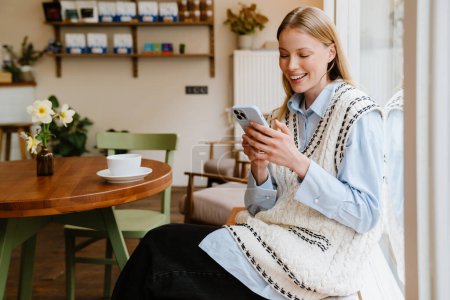 Foto de Joven hermosa mujer sonriente en camisa y chaleco con bolsa grande sosteniendo y usando su teléfono mientras está sentada en la acogedora cafetería - Imagen libre de derechos