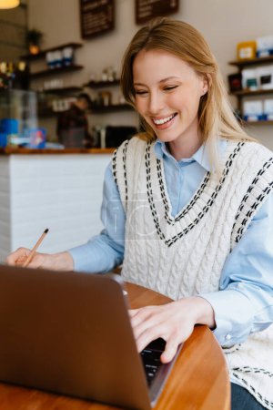 Téléchargez les photos : Young beautiful long-haired smiling happy woman in shirt and vest with pencil in her hand working with laptop while sitting in cafe - en image libre de droit