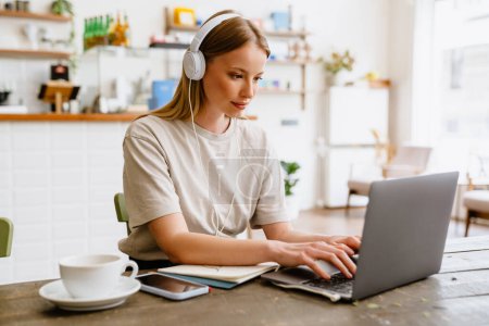 Foto de Joven hermosa mujer tranquila de pelo largo en los auriculares que trabajan con el ordenador portátil, mientras está sentado en la acogedora cafetería - Imagen libre de derechos