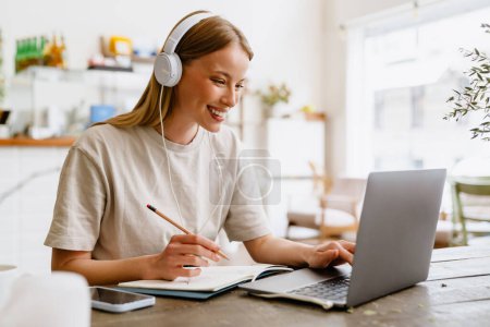 Foto de Joven hermosa de pelo largo sonriente mujer feliz en los auriculares haciendo su tarea con lápiz y cuaderno de escribir en el ordenador portátil mientras está sentado en la cafetería - Imagen libre de derechos