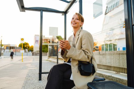 Photo for Young white woman smiling and using cellphone while sitting at bus station - Royalty Free Image