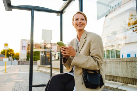 Photo for Young white woman smiling and using cellphone while sitting at bus station - Royalty Free Image