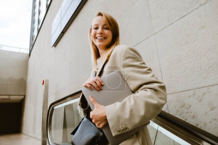 Téléchargez les photos : Jeune femme blanche souriant et tenant ordinateur portable tout en se tenant debout sur l'escalator à l'extérieur - en image libre de droit