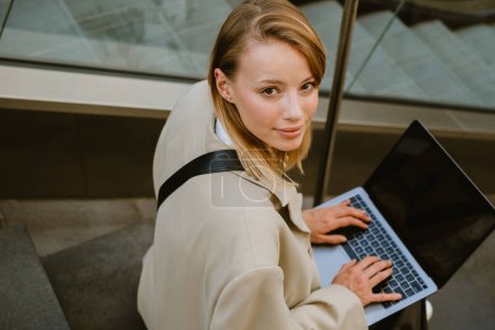 Photo for Young white woman smiling and using laptop while sitting on stairs outdoors - Royalty Free Image