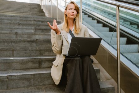 Téléchargez les photos : Jeune femme sérieuse blanche utilisant un ordinateur portable tout en étant assis sur les escaliers à l'extérieur - en image libre de droit