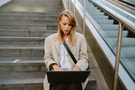 Photo for Young white serious woman using laptop while sitting on stairs outdoors - Royalty Free Image