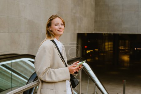 Foto de Young white woman smiling and using cellphone while standing by escalator outdoors - Imagen libre de derechos