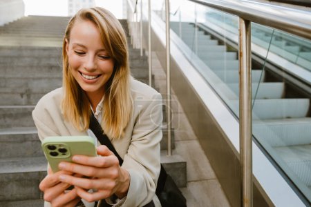 Photo for Young white woman smiling and using cellphone while sitting on stairs outdoors - Royalty Free Image