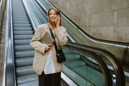 Téléchargez les photos : Jeune femme blanche parlant sur un téléphone portable et tenant un ordinateur portable tout en descendant l'escalator à l'extérieur - en image libre de droit