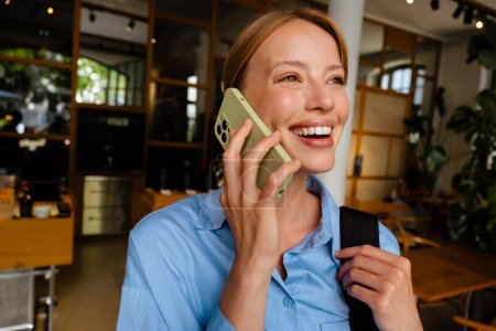 Photo for Young white woman wearing shirt smiling while talking on cellphone indoors - Royalty Free Image