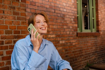 Téléchargez les photos : Blanc jeune femme souriant et parlant sur téléphone portable tout en se reposant dans le café à l'extérieur - en image libre de droit